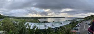 Panoramic view of the roof of a building, cars in the parking lot, and water in the background and clouds across the sky.